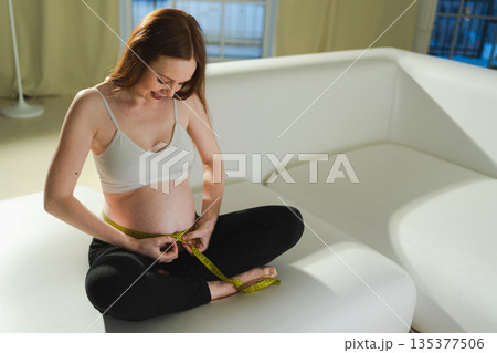 Pregnancy motherhood. Pregnant woman measuring big belly. Girl measuring her belly with tape to keep track of her fetus development. Woman enjoying pregnancy. Maternity tenderness parenthood new life Pregnancy motherhood. Pregnant woman measuring big belly. Girl measuring her belly with tape to keep track of her fetus development. Woman enjoying pregnancy. Maternity tenderness parenthood new life 135377506