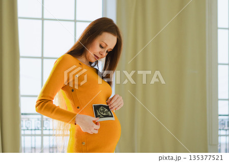 Prenatal ultrasound screening. Pregnant woman holding sonogram picture of unborn baby inside her big belly. Happy expectant mom with fetus usi scan image. Young mother enjoying pregnancy. Maternity Prenatal ultrasound screening. Pregnant woman holding sonogram picture of unborn baby inside her big belly. Happy expectant mom with fetus usi scan image. Young mother enjoying pregnancy. Maternity 135377521