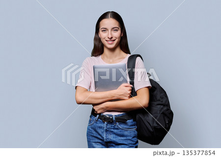 Portrait of beautiful female teenage student with backpack, laptop on gray background Portrait of beautiful female teenage student with backpack, laptop on gray background 135377854