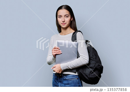 Portrait of beautiful female teenage student with backpack, laptop on gray background 135377858
