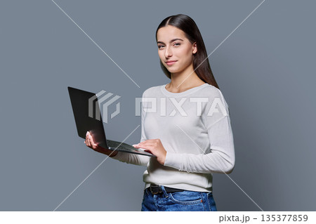 Portrait of smiling teenage girl holding laptop on gray studio background 135377859