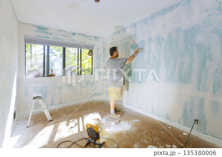 Worker is applying putty plaster to walls of room with windows surrounded by tools dust. Worker is applying putty plaster to walls of room with windows surrounded by tools dust. 135380006