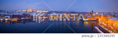 High resolution winter panorama of Prague featuring Charles Bridge, Prague Castle and the Vltava River at dusk. Snow covered historic city illuminated by warm lights creates a magical European winter 135380295
