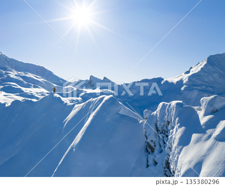 Freeride skier standing on a snowy mountain ridge, preparing for descent in pristine alpine terrain. Bright winter sun, dramatic peaks and untouched snow create a powerful adventure atmosphere. 135380296