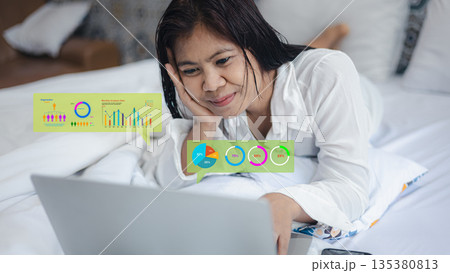 Happy Asian businesswoman lying on bed using laptop with holographic financial data charts. Concept of smart remote work, online trading, and digital business analysis success. 135380813