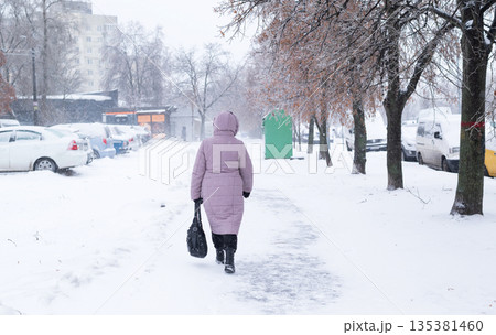 A woman in a pink winter coat walks down a snow-covered city sidewalk during a heavy snowfall. 135381460