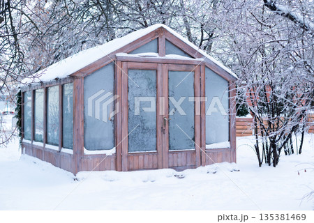 A wooden garden greenhouse covered in snow and frost, surrounded by icy winter trees in a backyard 135381469