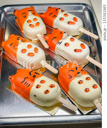 Cute fox-shaped cakesicles with orange and white chocolate glaze on a metal tray in a bakery shop Cute fox-shaped cakesicles with orange and white chocolate glaze on a metal tray in a bakery shop 135381865