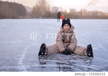 A happy girl wearing ice skates sits on the surface of a frozen lake while her father and sister skate 135382112