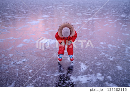 A tired child while skating. Reflection of the red setting sun on the ice of the frozen sun in the late evening A tired child while skating. Reflection of the red setting sun on the ice of the frozen sun in the late evening 135382116