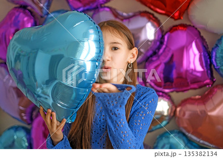 The girl blows a kiss, holding a balloon heart, Congratulations on Valentine's Day. The wall is decorated with bright colorful foil balloons The girl blows a kiss, holding a balloon heart, Congratulations on Valentine's Day. The wall is decorated with bright colorful foil balloons 135382134