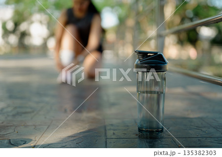 Water bottle in focus as female runner adjusts shoe during outdoor workout break in park Water bottle in focus as female runner adjusts shoe during outdoor workout break in park 135382303