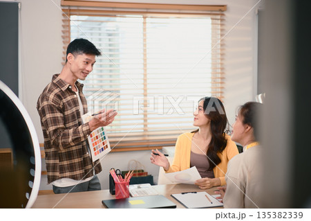 Fashion design team discussing clothing details and color selection at studio, reviewing samples and planning next collection together 135382339