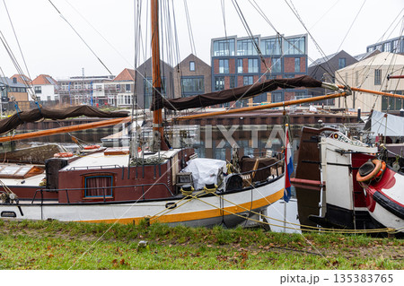 Moored Dutch sailing boats with wooden masts and furled sails line a canal beside modern glass buildings and red tiled roofs. 135383765