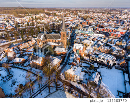 Snow covered town unfolds around a central church with tall steeple, dense rooftops, and winding streets, framed by wooded edges and open fields in crisp winter light. 135383770