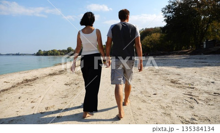 Romantic couple holding hands of each other and walking at coastline. Happy pair going along seashore at sunny day. Lovers spending time together at resort. Concept of vacation or holiday. Slow motion Romantic couple holding hands of each other and walking at coastline. Happy pair going along seashore at sunny day. Lovers spending time together at resort. Concept of vacation or holiday. Slow motion 135384134