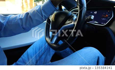 Male hand of businessman holds steering wheel while driving electrical vehicle at city. Businessperson operating electric car on urban road. Young man commuting to work at sunny day. Close up 135384141