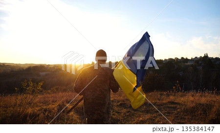 Young man in military uniform jogs with waving flag of Ukraine at sunset. Male soldier of ukrainian army running with lifted blue-yellow banner at countryside. Concept of invasion resistance 135384147