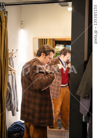 Elegant man trying on wool vintage blazer in fitting room, wearing clothing in front of mirror. 135385155