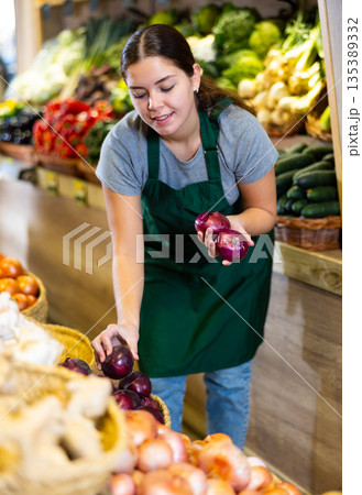 Female grocery store worker arranges red onion and other vegetables on counter and display case Female grocery store worker arranges red onion and other vegetables on counter and display case 135389332