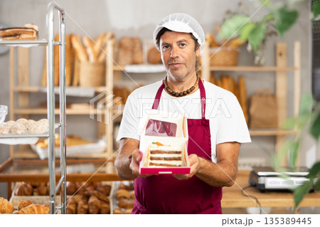 Middle-aged salesman offering a piece of cake packed in box in bakery Middle-aged salesman offering a piece of cake packed in box in bakery 135389445