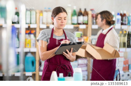 Girl seller, with tablet in hands, make inventory in store. Man employee in background 135389613