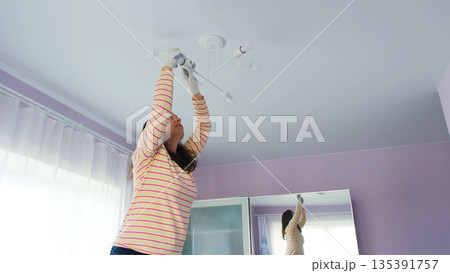 A woman stands on a stepladder screwing a new light bulb into a ceiling lamp in a room with purple walls and natural light A woman stands on a stepladder screwing a new light bulb into a ceiling lamp in a room with purple walls and natural light 135391757