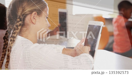 Schoolgirl sitting at school desk viewing code overlay on tablet computer, with blurred classmate 135392573