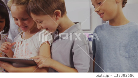 Girl in white floral top holding tablet with peers tapping tablet in classroom, desks whiteboard 135392586