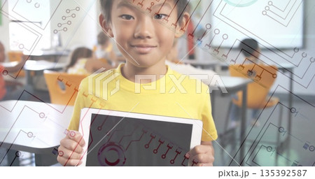 Standing boy holding tablet displaying circuit-pattern graphics in classroom, with orange seats 135392587