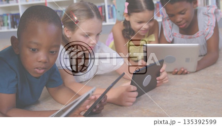 Lying four children wearing colorful clothes using tablets in reading area, with bookshelves behind 135392599