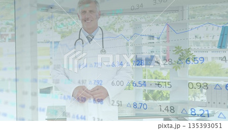 Standing male doctor wearing white lab coat smiling in office, with stethoscope and data overlays 135393051