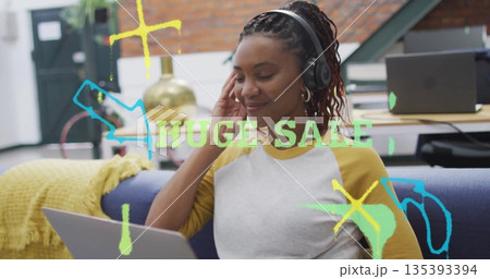 Typing Black woman wearing headphones on couch in office, working on laptop with neon sale text 135393394