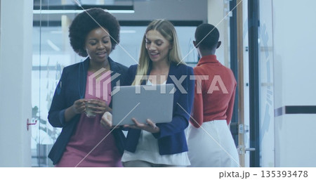 Two women walking and examining laptop in corridor with frosted partitions, holding glass of water 135393478