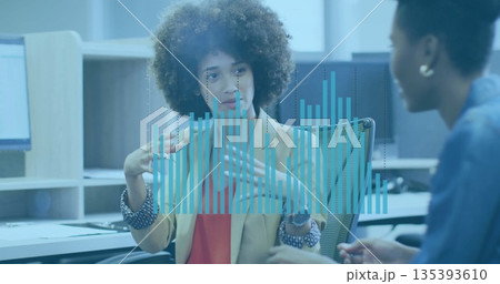 Gesturing curly haired woman wearing blazer explaining data overlay in office, with monitors Gesturing curly haired woman wearing blazer explaining data overlay in office, with monitors 135393610