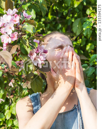Young woman sneezes in the park against the background of a flowering tree. Allergy to pollen concept. Young woman sneezes in the park against the background of a flowering tree. Allergy to pollen concept. 135394175