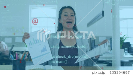 Presenting Korean woman wearing grey blazer displaying charts at office desk, with monitors 135394697