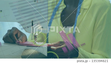 Seamstress guiding pink fabric under sewing machine in studio, with measuring tape and charts Seamstress guiding pink fabric under sewing machine in studio, with measuring tape and charts 135394821