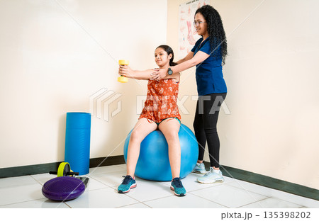 Physiotherapist helping patient woman with dumbbell sitting on rehabilitation ball. Chiropractor helping woman patient with dumbbell sitting on Fitness Ball Physiotherapist helping patient woman with dumbbell sitting on rehabilitation ball. Chiropractor helping woman patient with dumbbell sitting on Fitness Ball 135398202