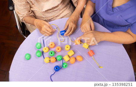 Caregiver and senior woman playing stringing beads game for dementia prevention 135398251