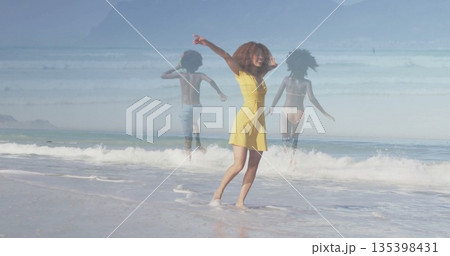 Smiling mother wearing yellow sundress raising arms at sandy shoreline, children running into surf 135398431