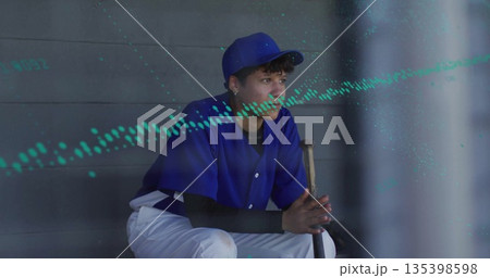 Sitting teen in blue uniform and cap gripping wooden bat on dugout bench, gazing toward field 135398598