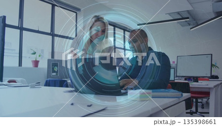 Two office professionals collaborating on laptop in open-plan office, with papers and plant 135398661