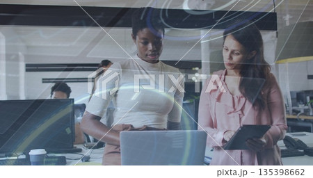 Collaborating women wearing office attire using overlays on laptop, tablet in office, with monitors 135398662