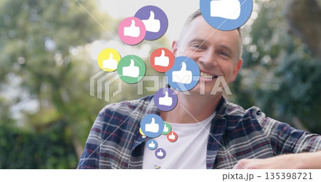 Smiling man wearing plaid shirt sitting in backyard, with floating thumbs-up icons, copy space 135398721