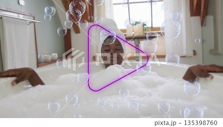 Woman with towel-wrapped hair soaking in foam-filled white bathtub in bathroom, with neon play icon Woman with towel-wrapped hair soaking in foam-filled white bathtub in bathroom, with neon play icon 135398760