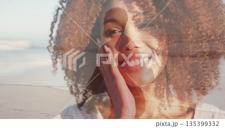 Smiling woman in white blouse touching cheek on beach, with man in coat walking silhouette Smiling woman in white blouse touching cheek on beach, with man in coat walking silhouette 135399332