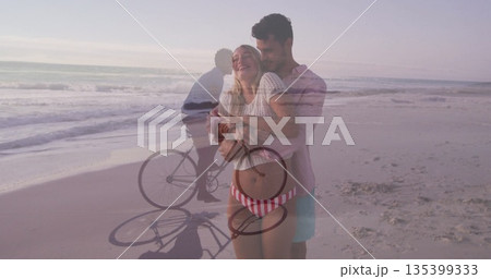 Embracing pregnant couple wearing striped bikini bottom on sandy beach, with bicycle silhouette 135399333