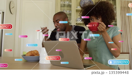 Man and woman eating and drinking at home kitchen table, with laptop, red mug and notifications 135399448
