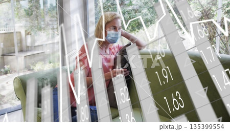 Holding pillow, woman wearing surgical mask studying financial chart overlay on green sofa at home Holding pillow, woman wearing surgical mask studying financial chart overlay on green sofa at home 135399554